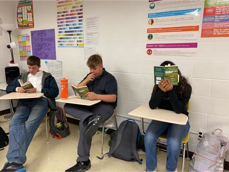 three students sit in desks and read books  
